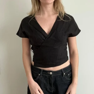 Svart omlottopp med knyt - Snygg svart omlottopp med korta ärmar och knytning i ryggen. Toppen har en v-ringad design och är croppad för en modern look. Perfekt för att skapa en stilren outfit.