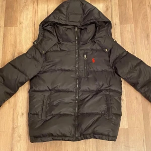 Svart Ralph lauren Jacka - Säljer en svart dunjacka från Ralph Lauren med huva och dragkedja. Jackan har en röd logotyp på bröstet och är perfekt för kyliga dagar