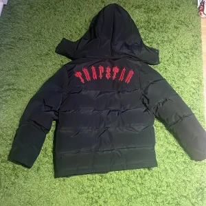 Svart pufferjacka från Trapstar - Säljer en svart pufferjacka från Trapstar med röd broderad text på ryggen och en cool logga på bröstet. Jackan har en dragkedja framtill och en huva för extra skydd. Perfekt för att hålla sig varm och stilren under kyliga dagar.