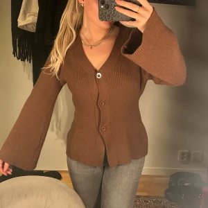 Brun ribbad kofta med knappar - Säljer en mysig brun kofta med ribbad struktur och knappar framtill. Koftan har långa ärmar och en v-ringad design, perfekt för kyligare dagar.