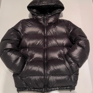 Svart pufferjacka Zara - Säljer en svart pufferjacka från Zara med huva och dragkedja. Jackan är perfekt för kalla dagar och har en glansig yta som ger en modern look. Den är designad för att hålla dig varm och bekväm. Ord pris 1400kr. Skicket är 9/10 Inga skador eller skavanker