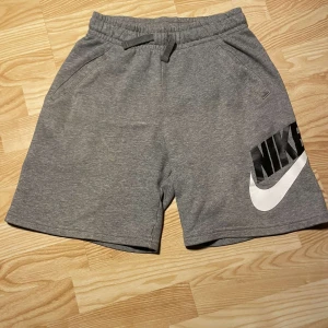 Nike shorts grå - Snygga grå shorts från Nike med elastisk midja och snörning. De har en stor Nike-logga i svart och vitt på ena benet. Perfekta för en avslappnad stil.