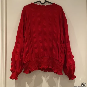 Röd långärmad tröja med volangdetaljer - Säljer en snygg röd tröja med långa ärmar och volangdetaljer vid ärmslut och nederkant. Tröjan har en unik textur och en knappdetalj i nacken. Perfekt för att liva upp garderoben!