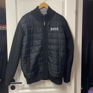 Svart jacka från Boss - Snygg svart jacka från Boss med quiltad framsida och dragkedja. Insidan har ett coolt camouflagemönster i svart och vitt. Perfekt för kyligare dagar med sin långa ärmar och ribbade detaljer.