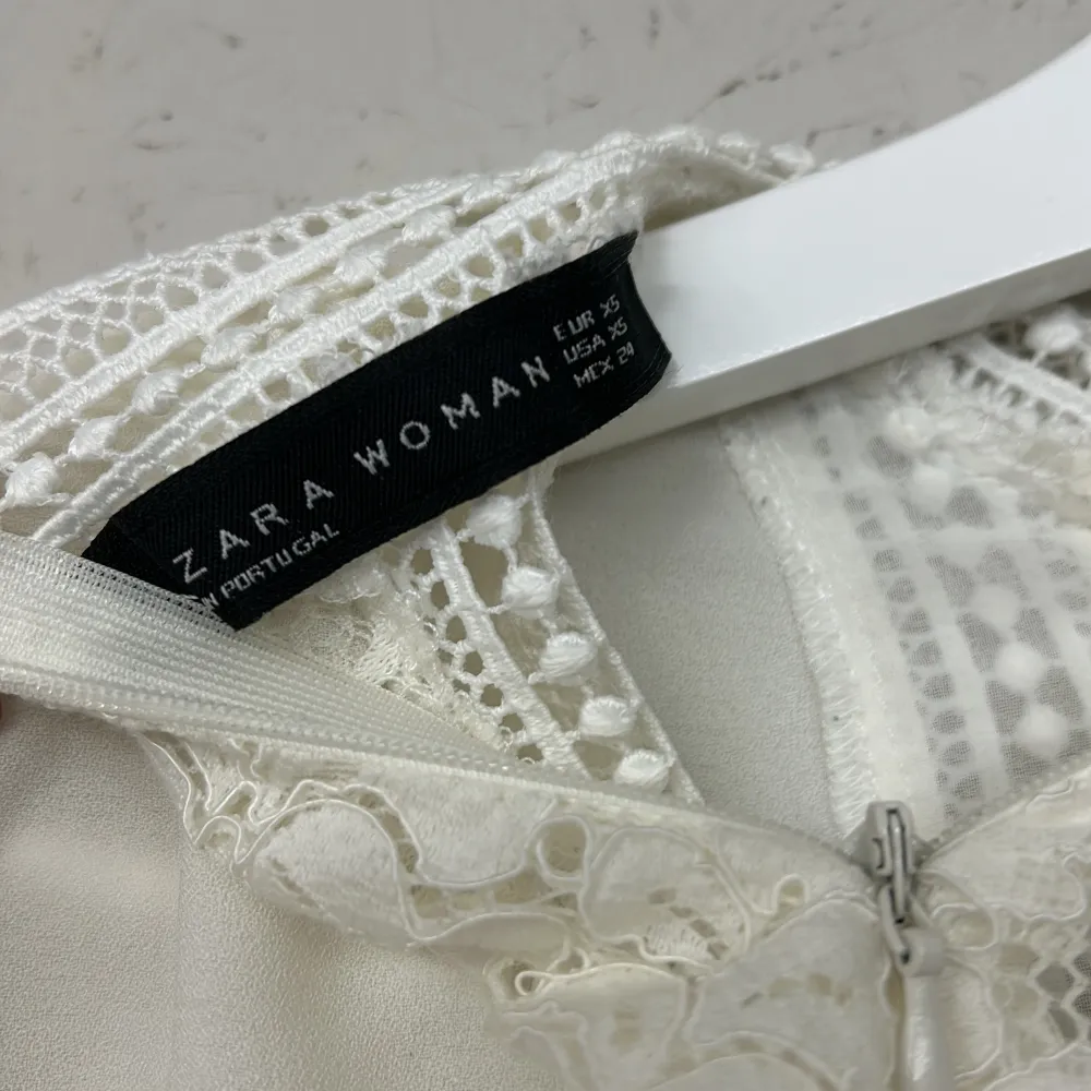 Elegant vit blus från Zara med vackra spetsdetaljer och korta ärmar. Blusen har en fin volangkant och en diskret dragkedja i ryggen. Perfekt för en stilren look.. Puserot.