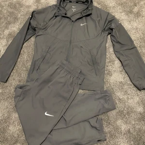 Nike tracksuit - Säljer en grå tränings tracksuit från Nike med dragkedja och huva. Jackan har långa ärmar och är perfekt för löpning. Den är tillverkad i ett syntetmaterial som är lätt och bekvämt. Nike-loggan syns på bröstet och ärmen.