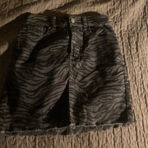 Zebramönstrad jeanskjol - Snygg zebramönstrad jeanskjol i svart och grått. Kjolen har en rå kant nertill och klassisk femficksdesign med knapp och dragkedja framtill. Perfekt för en trendig look!