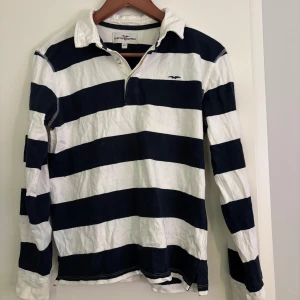 Randig pikétröja från Ralph Lauren - Säljer en klassisk randig pikétröja från Hampton Republic i marinblått och vitt. Tröjan har långa ärmar och en krage med knappslå. Perfekt för en avslappnad och stilren look.