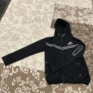 Nike tech  - Säljer en svart jacka från Nike med dragkedja och huva. Jackan har långa ärmar och en stilren design med vita detaljer. Perfekt för en sportig look. INTE ÄKTA! 