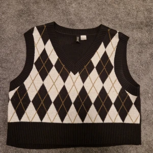 Svart och vit rutig väst från H&M Divided - Snygg svart och vit väst från H&M Divided med ett klassiskt rutigt mönster. Västen har en v-ringad design och ribbade kanter. Perfekt för att lägga till en stilren touch till din outfit.