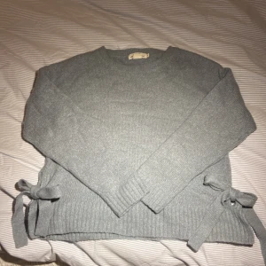 Grå stickad tröja med knyten på sidorna - Mysig grå stickad tröja med långa ärmar och dekorativa knytband på sidorna.ribbade nederkant och ärmslut. En stilren och bekväm tröja för alla tillfällen.nopp fri 