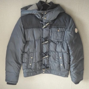 Grå/grå dunjacka med knäppning - Säljer en snygg grå/blå dunjacka från Moncler med unik knäppning framtill. Jackan har en praktisk huva och flera fickor med knappar. Perfekt för kyliga dagar! Har mått om de skulle behövas!!