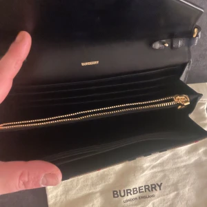 Rutig plånbok från Burberry - Snygg rutig plånbok från Burberry med klassiskt mönster i beige, svart och rött. Plånboken har en praktisk dragkedja och en elegant design med en svart klaff. Perfekt för att hålla ordning på kort och kontanter.