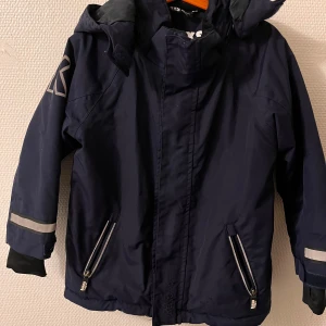 Mörkblå jacka från Kaxs - Säljer en mörkblå jacka från Kaxs med huva och reflexdetaljer på ärmarna. Jackan har dragkedja framtill och två fickor med dragkedjor. Perfekt för kyliga dagar och håller dig varm och synlig. 🧥✨