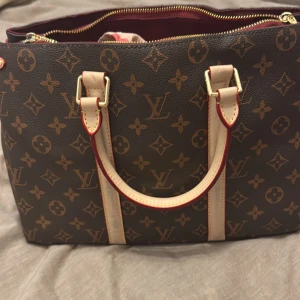 Brun monogram handväska  - Säljer en elegant brun handväska med monogrammönstret. Väskan har ljusa handtag med röda kanter och en dragkedja upptill. Insidan är klädd i röd textil och erbjuder gott om utrymme för dina tillhörigheter. Skydds tejp är kvar så oanvänd såklart. 