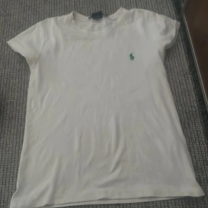 Vit t-shirt från Ralph Lauren - Säljer en klassisk vit t-shirt från Ralph Lauren med en grön broderad logotyp på bröstet. T-shirten har en normal passform och korta ärmar, perfekt för en avslappnad stil.