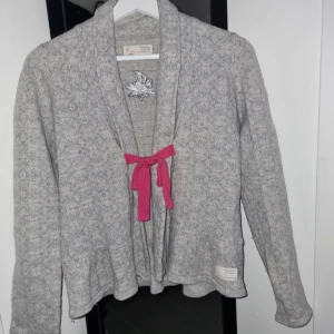 Grå kofta från Odd Molly - Säljer en mysig grå kofta från Odd Molly med en rosa rosett framtill. Koftan har ett broderat mönster på ryggen och långa ärmar. 