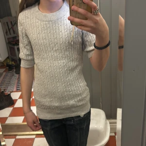 Grå stickad tröja - Säljer en stilren grå stickad tröja med kabelstickat mönster. Tröjan har korta ärmar och en rund halsringning. Perfekt för en avslappnad och bekväm stil. Passar bra till jeans eller kjol.