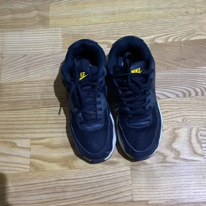 Svarta Nike Air Max sneakers - Säljer ett par svarta Nike Air Max sneakers med synlig luftdämpning i sulan. Skorna har en klassisk design med snörning och gula detaljer på tungan. Perfekta för en sportig look.