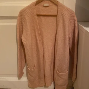 Ljusrosa kofta från By Malene Birger - Säljer en elegant Ljusrosa kofta från By Malene Birger. Den är tillverkad i en mjuk blandning av alpacka, ull och polyamid, vilket gör den både varm och bekväm. Koftan har långa ärmar och praktiska fickor framtill. Perfekt för kyliga dagar!