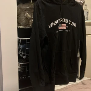 Svart hoodie från Vins Polo Club - Säljer en svart hoodie från Vins Polo Club med tryck på framsidan. Tröjan har en klassisk design med lång ärm och en praktisk ficka framtill. Perfekt för en avslappnad stil.