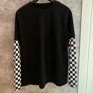 Svart tröja med schackrutiga ärmar - Säljer en cool svart tröja med långärmade schackrutiga detaljer i svart och vitt. 