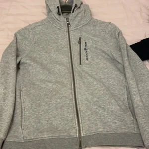 Grå huvtröja från Sail Racing - Säljer en grå huvtröja från Sail Racing med dragkedja framtill och en bröstficka med logotyp. Tröjan har långa ärmar och en bekväm passform. Perfekt för en avslappnad stil. Pris kan diskuteras via köp! Storlek 160