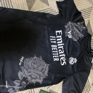 Svart fotbollströja från Adidas - Snygg svart fotbollströja från Adidas med tryck av stora grå rosor och texten 'Emirates Fly Better'. Tröjan har korta ärmar och en stilren design med klubbmärke och Y-3 logga. Perfekt för fotbollsfans som vill ha något unikt.