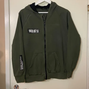 Grön huvtröja från Woxo - Säljer en grön huvtröja från Woxo med tryck på bröstet och ryggen. Tröjan har dragkedja framtill och en bekväm huva. För barn i storlek 170