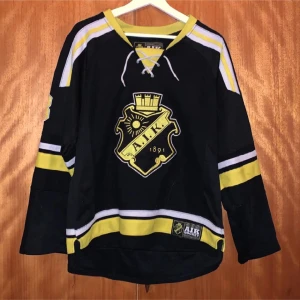 Svart AIK hockeytröja - Säljer en svart AIK hockeytröja med gula och vita detaljer. Tröjan har långa ärmar och ett stort AIK-emblem på framsidan. 