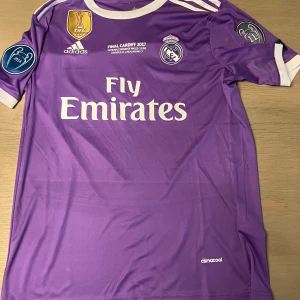 Lila Real Madrid fotbollströja - Säljer en lila Real Madrid fotbollströja från Adidas med 'Fly Emirates' tryck på framsidan. Tröjan har korta ärmar och är dekorerad med märken för FIFA och UEFA. På baksidan finns namnet 'Ronaldo' och nummer 7. Perfekt för fans av laget!