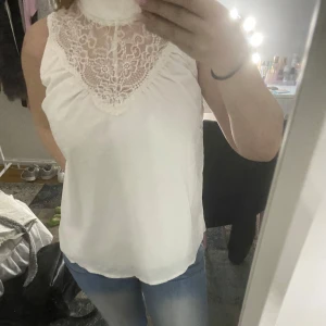 Vit ärmlös blus med spetsdetaljer - Säljer en elegant vit blus med spetsdetaljer vid halsen. Blusen är ärmlös och har en luftig passform, perfekt för varmare dagar. Den har en hög hals med vackra spetsmönster som ger en romantisk touch.