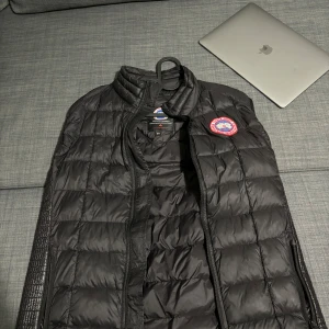 Svart dunväst från Canada Goose - Säljer en stilren svart dunväst från Canada Goose. Västen har en quiltad design och en dragkedja framtill. Perfekt för kyligare dagar och ger en snygg look. Passar bra till både vardag och utomhusaktiviteter.