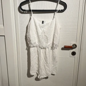 Vit jumpsuit med spetsdetaljer - Säljer en söt vit jumpsuit från Divided med tunna axelband och vackra spetsdetaljer framtill. Perfekt för sommarens varma dagar. Jumpsuiten har en avslappnad passform och är tillverkad i ett lätt material.