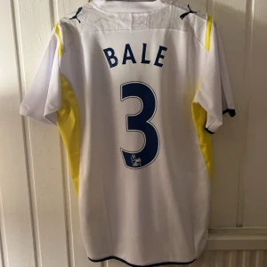Bale Tottenham fotbollströja - Säljer en vit fotbollströja med gula och svarta detaljer från Puma. Tröjan har korta ärmar och ett tryck med 'Bale' och nummer 3 på baksidan. 