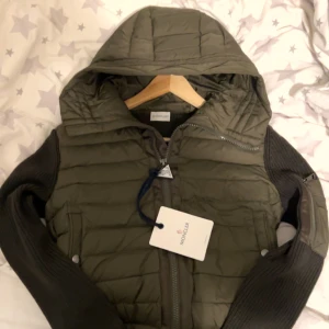 Grön quiltad jacka från Moncler - (Har kvitto)Snygg grön quiltad jacka från Moncler med huva och dragkedja. Jackan har stickade ärmar i mörkare ton och en broderad Moncler-logga på ärmen. Perfekt för kyligare dagar.