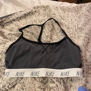 Grå sporttopp från Nike - Säljer en grå sporttopp från Nike med svarta detaljer och elastiskt band med Nike-logga. Toppen har en brottarrygg och är perfekt för träning. Den är tillverkad i ett mjukt och bekvämt material. Står att det ska vara en S men skulle säga att det mer är en xs🤩
