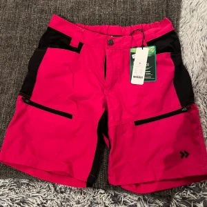 Rosa och svarta shorts med dragkedjor - Snygga rosa shorts med svarta detaljer och praktiska dragkedjor på fickorna. Perfekta för en aktiv dag utomhus. Friluftsshorts, ny pris 300kr  