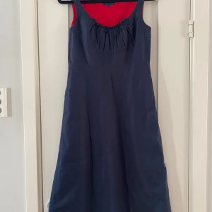 Mörkblå klänning från Tommy Hilfiger strl 36 - Snygg mörkblå klänning från Tommy Hilfiger med röd insida. Klänningen har en elegant rynkad detalj vid bysten och är ärmlös. Perfekt för en stilren look. Ny marinblå klänning i strl 4 (36) från Tommy Hilfiger.  Ny, aldrig använd.  Klänningen har en underklänning och knäpps med dragkedja i sidan.  Mått Över bröstet: ca 41 cm Längd från axeln och ner: ca 101 cm  100% bomull  Kommer från ett djur och rökfritt hem.