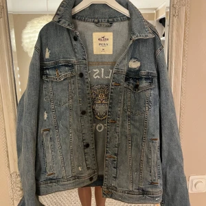 Jeansjacka från Hollister - Säljer en cool jeansjacka från Hollister med ett stort broderat tigerhuvud och texten 'Hollister California' på ryggen. Jackan har en sliten look med flera revor och knappar framtill. 