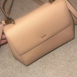 Beige axelväska från DKNY - Snygg beige axelväska från DKNY i skinn med justerbar axelrem och stilren design. Väskan har en praktisk storlek och är perfekt för att bära dina dagliga nödvändigheter. Den har en diskret logotyp på framsidan och metallfötter på undersidan för extra skydd. Väldigt oanvänd då jag bara använt den ungefär 2 gånger.