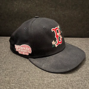 Boston red socks keps från New Era - Snygg svart keps från New Era med broderad boston red socks med blommotiv på framsidan. Kepsen har även en World Series-märke på sidan och MLB-logga baktill. Perfekt för sportfantaster! Priset går att diskutera och det går att mötas upp i Stockholms området.