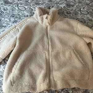 Beige teddyjacka - Mysig beige teddyjacka med dragkedja framtill och hög krage. Perfekt för kyliga dagar med sin fluffiga och varma design. 