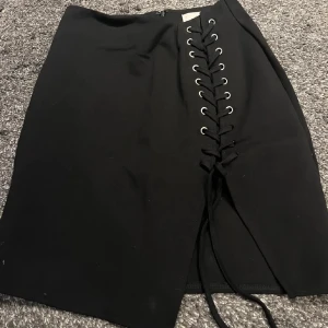 Svart kjol med snörning från Rebecca Stella - Snygg svart kjol från Rebecca Stella med cool snörning framtill och en diskret dragkedja i sidan. Perfekt för att ge en edgy touch till din outfit.