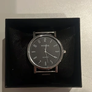 Svart klocka från Modiya - Stilren klocka från Modiya med svart urtavla och silverfärgade detaljer. Klockan har en klassisk design med tydliga siffror och kvartsurverk. Perfekt för en minimalistisk stil.
