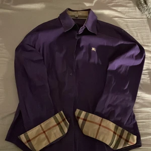 Lila skjorta från Burberry - Snygg lila skjorta från Burberry med klassiskt rutigt mönster på insidan av kragen och ärmsluten. Skjortan har långa ärmar och en liten broderad logga på bröstet. Perfekt för en stilren look. Skjortan är i XXS och passar folk från 140-160. Väldigt sparsamt använd