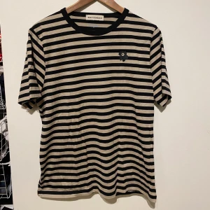 Randig t-shirt från Marimekko - Säljer en snygg randig t-shirt från Marimekko i svart och beige. Den har korta ärmar och ett litet blommönster på bröstet. Perfekt för en avslappnad stil. 🌼