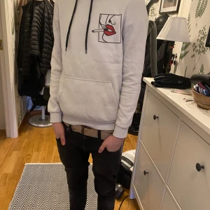 Vit hoodie från Pier One med konstnärligt tryck - Säljer en vit hoodie från Pier One med ett unikt konstnärligt tryck av ansikten i svart och rött på både fram- och baksidan. Hoodien har en klassisk passform med långa ärmar och en justerbar huva med svarta snören. Perfekt för en stilren och modern look.