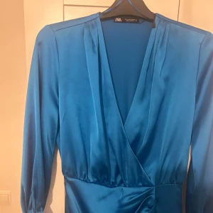 Blå omlottklänning från Zara - Snygg blå omlottklänning från Zara med långa ärmar och v-ringad design. Perfekt för en stilren look. Tillverkad i ett mjukt och glansigt material som ger en elegant känsla.