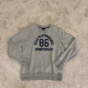 Grå sweatshirt från Peak Performance - Säljer en grå sweatshirt från Peak Performance med tryck '86 Sportswear' i marinblått på framsidan. Passar både män och kvinnor alltså unisex. Tröjan har långa ärmar och ribbade muddar vid ärmslut och nederkant. Perfekt för en avslappnad stil.
